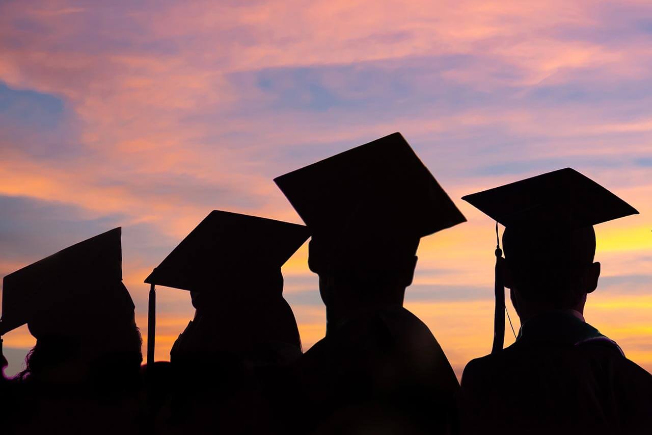 graduate silhouettes with graduation caps on looking at a sunset